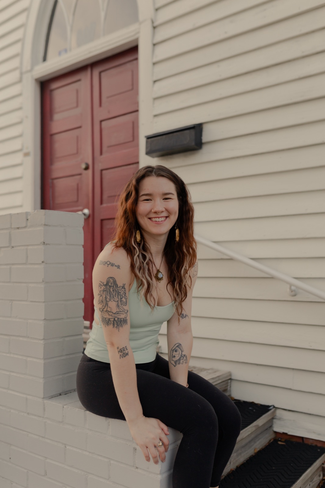 The maker behind Sidequest Sis, sitting on the front steps of a white building with a red door